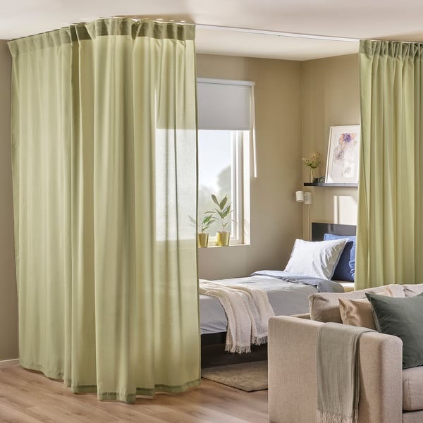 Cortinas verde-oliva dividem o quarto aconchegante com cortinas suaves. Cama e sofá visíveis.