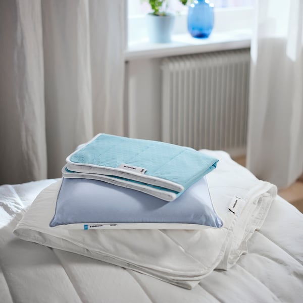 Uma cama bem arrumada com lençóis brancos e acessórios de cama azul claro, incluindo protetor de colchão, fronha e edredom dobrado.