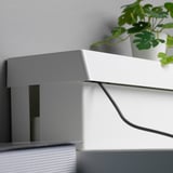 Caixa branca, provavelmente o SÄTTING, ocultando cabos. Tampa projetada para fácil acesso ao cabo. Planta próxima, cabo visível.