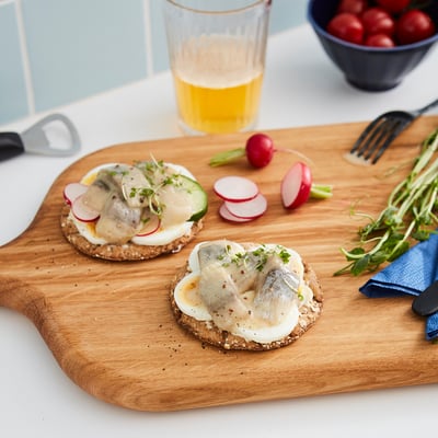 A tábua de madeira contém dois sanduíches abertos cobertos com arenque, legumes fatiados e verduras. Copo de bebida amarela, faca e rabanetes nas proximidades.