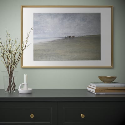 Quadro costeiro com moldura dourada na parede da hortelã sobre armário preto com alças douradas, livros, flores, vela.