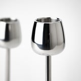 Dois castiçais de aço inoxidável elegantes e brilhantes com superfícies polidas e linhas curvas suaves. Um em primeiro plano, outro borrado no fundo.