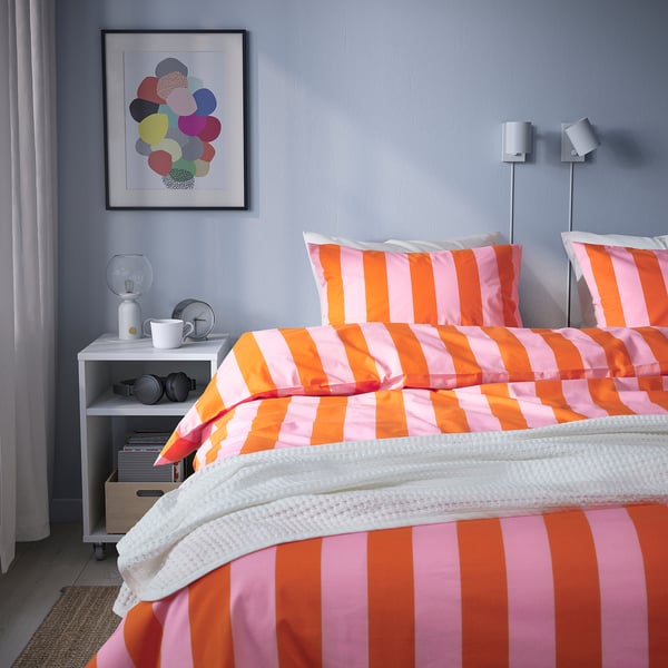 Quarto luminoso com roupa de cama listrada grande, arte colorida e uma pequena mesa de cabeceira.