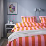Quarto luminoso com roupa de cama listrada grande, arte colorida e uma pequena mesa de cabeceira.