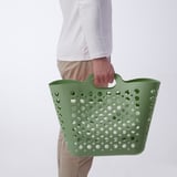 Pessoa segurando cesto de roupa suja de plástico flexível verde com alças, perfeito para transportar roupas.