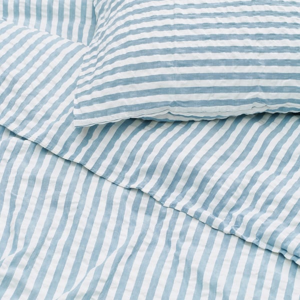Mostra roupa de cama listrada azul e branca com textura ondulada de sugador de seersucker, indicando tecido macio e respirável.