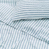 Mostra roupa de cama listrada azul e branca com textura ondulada de sugador de seersucker, indicando tecido macio e respirável.