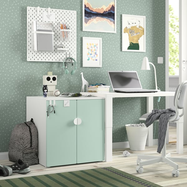 Pequeno armário SMÅSTAD verde e branco com gavetas, ao lado de uma secretária e cadeira. Design moderno, segurando vários itens pessoais.