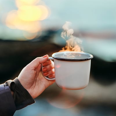 Mão a segurar uma caneca branca, fumegante, de esmalte.