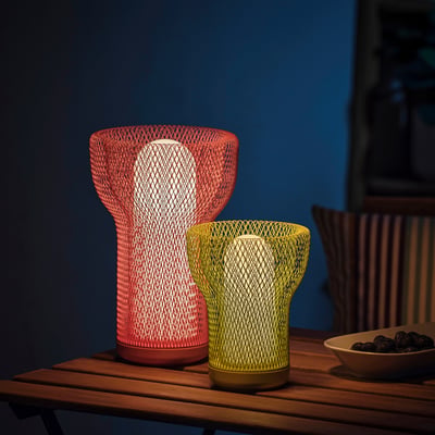 Duas lâmpadas SOLVINDEN ; vermelho e amarelo, sobre a mesa. Design de malha, uso interior/exterior. O vermelho é mais alto, tanto moderno como decorativo. Tigela de frutas ao lado.