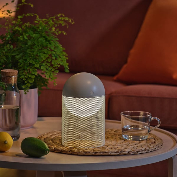 Uma lâmpada moderna em uma mesa, com uma planta, xícara e frutas por perto.