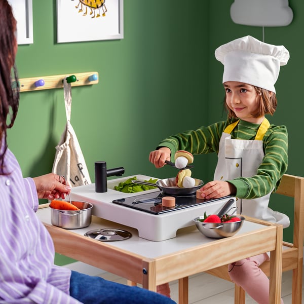 Criança no chapéu de chef cozinha no fogão de brinquedo com assistência de adulto.