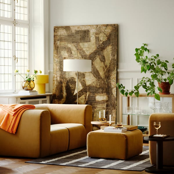 Sala de estar moderna com grande arte abstrata, sofá bronzeado, lâmpada amarela e plantas. O sofá é grande, quase enchendo a sala.