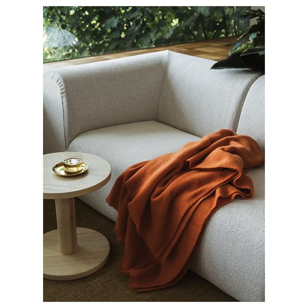 Sofá creme moderno com cobertor laranja, mesa lateral, vegetação.