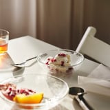 Tigela de vidro com design martelado contém sorvete coberto com sementes de romã, com copo vazio, garfo e colher em uma mesa branca.