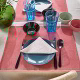 Um jogo americano rosa-vermelho com prato azul, copos e talheres pretos; protege a mesa e reduz o ruído.