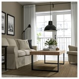 Sala de estar moderna com sofá bege, mesa de centro, luminária de chão e acesso à varanda. Grande janela, cortinas e decoração de plantas.