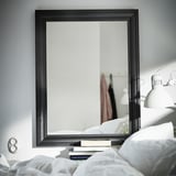 Espelho TOFTBYN preto na parede cinzenta, quarto refletor com cama, livros e candeeiro.