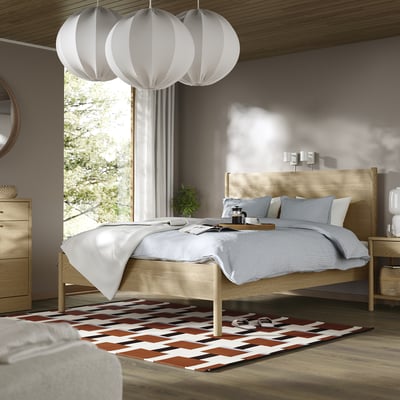 Cama minimalista com madeira clara, roupa de cama cinza, luminárias brancas, tapete colorido.