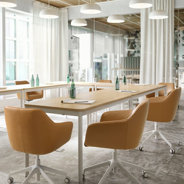 Escritório moderno com mesa de madeira clara, cadeiras giratórias TOSSBERG em couro bege, janelas grandes, candeeiros brancos.