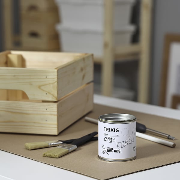 Uma pequena lata branca de tinta TRIXIG fica sobre papelão, ladeada por um pincel e uma chave de fenda. Caixas de madeira estão próximas, sugerindo projetos de bricolage.