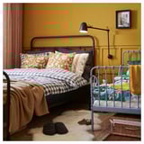 Quarto aconchegante com cama preta, travesseiros amarelos, cama infantil azul, tapete bege, chinelos, arandela de parede, quadro emoldurado nas paredes amarelas.