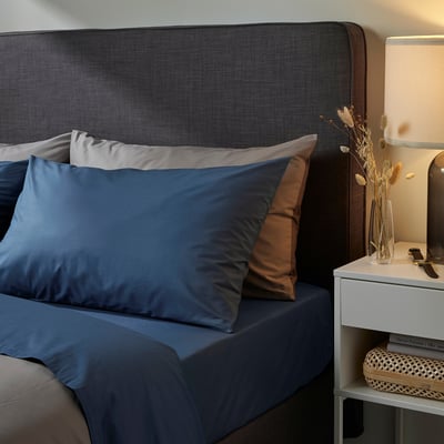 Quarto moderno com roupa de cama azul escuro, mesa lateral com abajur, livros e vaso com flores secas.