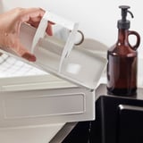 A mão segura o organizador de plástico inclinado, gotejamento de água, garrafa marrom com bomba no fundo. Mantém a cozinha arrumada.