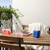 Mesa de pátio com cadeiras de madeira, alto-falante VAPPEBY, bebidas e plantas.