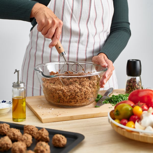 Pessoa mistura carne picada em tigela de vidro usando batedor de massa dinamarquesa. Azeite, ervas e tomates na mesa de madeira.