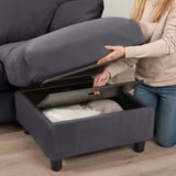 Uma pessoa abre um sofá otomano cinza para revelar um espaço de armazenamento cheio de travesseiros, mostrando seu design funcional.