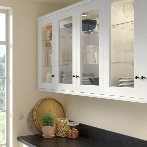 Modern white kitchen cabinet with glass doors, showcasing dishes and glasses, sitting above a dark countertop with a plant and a basket.