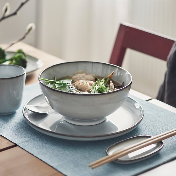Grey bowl on a plate with spoon and chopsticks, filled with soup and vegetables.