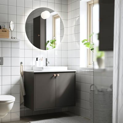 Modern minimalist bathroom: round mirror, white-tiled walls, grey HAVBÄCK vanity with sink, grey towel, plant, and striped laundry basket.