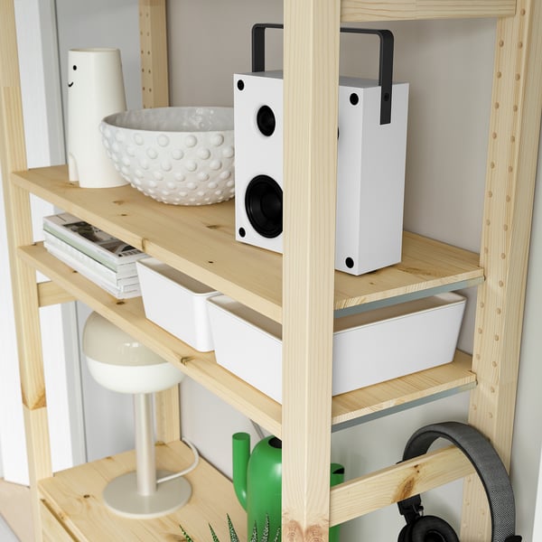 Wooden storage unit with shelves holding white speaker, white bowl, white containers, books, green watering can, and white lamp.