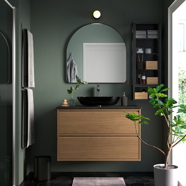 Modern bathroom with LINDBYN mirror. Arched, silver-framed mirror above wooden vanity with black sink, reflecting room.