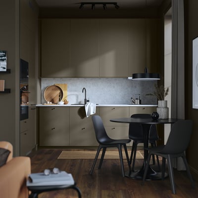 Modern kitchen with olive cabinetry, black chairs, and concrete backsplash. Sleek counters, pendant light, and wooden floor.