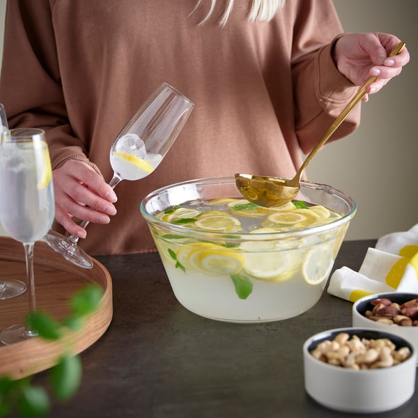 Person pours lemony drink from glass bowl using golden spoon. Bowl filled with lemon slices and mint. Other glasses, nuts, and napkins on table.