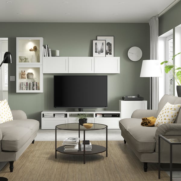 Living modern cu pereți verde salvie, televizor alb BESTÅ, două canapele mari bej, măsuță de cafea rotundă din sticlă și o lampă de podea elegantă.