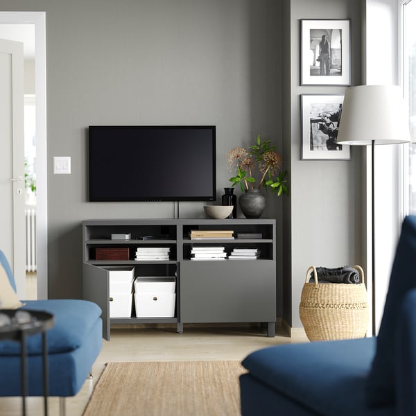 Living modern cu suport TV gri BESTÅ, televizor cu ecran plat, canapea albastră, coș țesut, lampă, cărți și fotografii alb-negru.