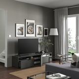 Living modern cu bancă TV gri BESTÅ, televizor negru, artă înrămată, lampă înaltă și măsuță de cafea din sticlă.