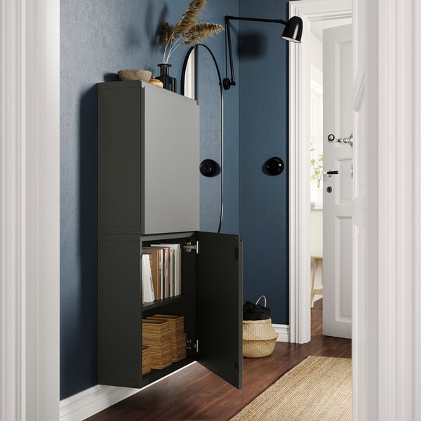 Living modern cu un dulap negru BESTÅ, care conține cărți și plante. Ușa deschisă arată cutii de depozitare și cărți. Lumină neagră montată pe perete.
