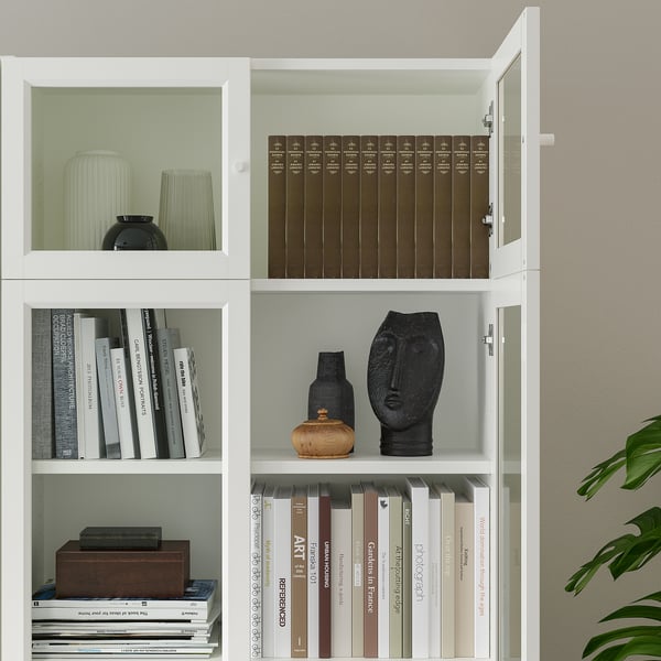 Bibliotecă albă cu rafturi, care expune cărți și obiecte decorative precum vaze și sculpturi. Uși din sticlă și panouri disponibile.