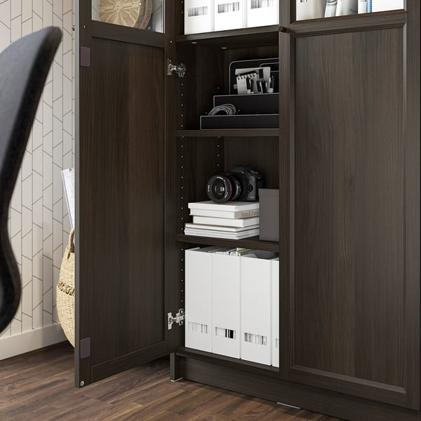 Dulap din lemn închis la culoare cu uși de sticlă deschise, dezvăluind cutii de depozitare, cărți și accesorii pentru camere bine organizate în interior.