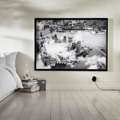 O fotografie mare înrămată alb-negru atârnă deasupra unui pat alb. Un teanc de cărți și o sticlă de sticlă stau pe podea lângă pat.