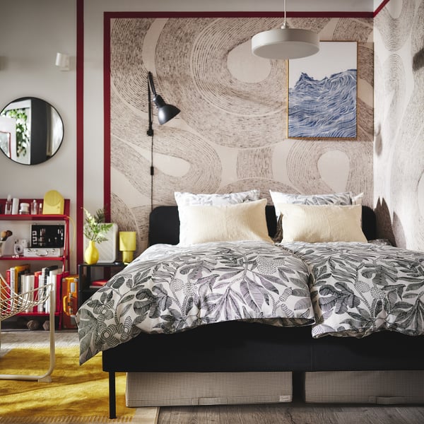 Dormitor modern cu lenjerie de pat florală neagră, două perne, artă de perete, lampă, măsuță laterală, oglindă, plante, cărți.