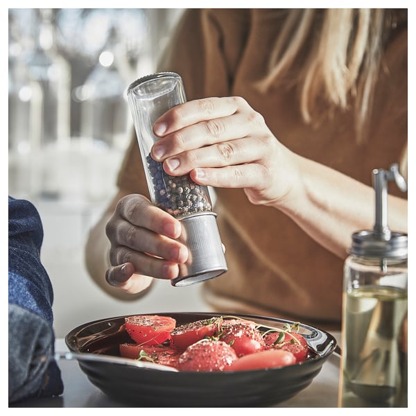 Persoana macină piperul peste farfurie cu râșnița de condimente CITRONHAJ. Sticlă transparentă, blat negru, capac din oțel inoxidabil. Masa are dozator de ulei verde, roșii.