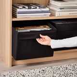 O persoană care scoate o cutie neagră din țesătură DRÖNA dintr-un raft din lemn, care conține diverse articole și este concepută pentru a maximiza spațiul de depozitare.