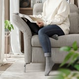 Persoană care citește carte pe canapea gri FAMMARP cu tetiere reglabile, purtând pulover alb, blugi și șosete gri.