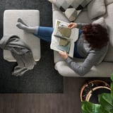 Persoana citește o carte pe canapea FAMMARP, gri, confortabilă, cu podea din lemn și plantă.